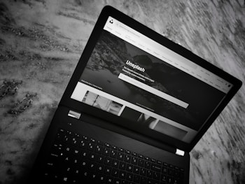 A black laptop is placed on a marble surface. The screen shows the homepage of the Unsplash website, with a header featuring navigation links and a search bar. The section below displays a large background image with text and several smaller images of photography content.