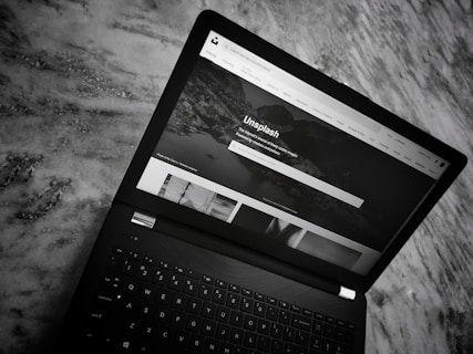 A black laptop is placed on a marble surface. The screen shows the homepage of the Unsplash website, with a header featuring navigation links and a search bar. The section below displays a large background image with text and several smaller images of photography content.