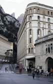 A historic building with arched windows and a stone facade stands prominently alongside a mountain landscape. There are multiple people walking along a road in front of the building. The area appears to be a cultural or tourist location, with signs and a museum entrance visible.