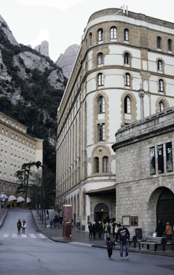 A historic building with arched windows and a stone facade stands prominently alongside a mountain landscape. There are multiple people walking along a road in front of the building. The area appears to be a cultural or tourist location, with signs and a museum entrance visible.
