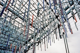 An intricate installation of hanging metallic rods suspended from a modern architectural ceiling. The structure consists of crisscrossing beams with reflective surfaces, creating a complex geometric pattern. The rods vary in color, with some displaying a shiny silver tone while others feature darker hues, such as blue and red.