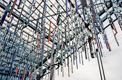 An intricate installation of hanging metallic rods suspended from a modern architectural ceiling. The structure consists of crisscrossing beams with reflective surfaces, creating a complex geometric pattern. The rods vary in color, with some displaying a shiny silver tone while others feature darker hues, such as blue and red.