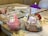 On a wooden table, there is a playful and artistic arrangement of two bowls of ice cream. One bowl has a character-themed scoop in pink, decorated with large candy eyes and a snout resembling a pig. The other bowl contains an elaborate sundae with vanilla and chocolate ice cream, topped with sliced bananas, strawberries, nuts, whipped cream, chocolate drizzle, and a wafer roll, garnished with a purple cocktail umbrella. In the background, a child’s hand is seen holding a colored pencil next to a coloring page.