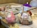 On a wooden table, there is a playful and artistic arrangement of two bowls of ice cream. One bowl has a character-themed scoop in pink, decorated with large candy eyes and a snout resembling a pig. The other bowl contains an elaborate sundae with vanilla and chocolate ice cream, topped with sliced bananas, strawberries, nuts, whipped cream, chocolate drizzle, and a wafer roll, garnished with a purple cocktail umbrella. In the background, a child’s hand is seen holding a colored pencil next to a coloring page.