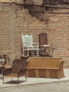 An outdoor scene featuring several wicker chairs and a white painted rocking chair arranged against a rustic brick wall. The chairs are stacked and positioned on top of a yellow wicker sofa, creating a display of vintage and eclectic furniture. The setting appears to be a back alley or courtyard with a raw, urban texture.
