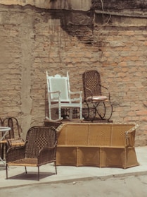 An outdoor scene featuring several wicker chairs and a white painted rocking chair arranged against a rustic brick wall. The chairs are stacked and positioned on top of a yellow wicker sofa, creating a display of vintage and eclectic furniture. The setting appears to be a back alley or courtyard with a raw, urban texture.