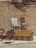An outdoor scene featuring several wicker chairs and a white painted rocking chair arranged against a rustic brick wall. The chairs are stacked and positioned on top of a yellow wicker sofa, creating a display of vintage and eclectic furniture. The setting appears to be a back alley or courtyard with a raw, urban texture.