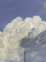 Towering white cumulus clouds dominate the scene against a backdrop of clear blue sky. A tall communication tower stands in the lower portion, dwarfed by the vast cloud formations. Several birds can be seen flying near the clouds.