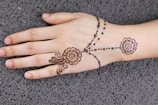 A hand resting on a textured surface, adorned with intricate henna designs. The artwork features floral patterns and delicate dots running across the hand, creating an elegant and traditional look.