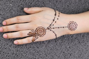 A hand resting on a textured surface, adorned with intricate henna designs. The artwork features floral patterns and delicate dots running across the hand, creating an elegant and traditional look.