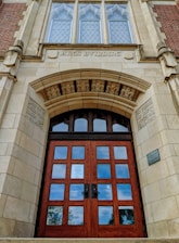 A grand, old architectural building entrance featuring a large wooden door with multiple panels and ornate stone carvings around the arch. Above the door are decorative windows with diamond-shaped glass panes, and the words 'Arts Building' etched into the stone. The structure is made of a combination of brick and stone, showcasing intricate detailing and craftsmanship.