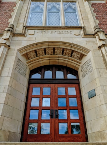 A grand, old architectural building entrance featuring a large wooden door with multiple panels and ornate stone carvings around the arch. Above the door are decorative windows with diamond-shaped glass panes, and the words 'Arts Building' etched into the stone. The structure is made of a combination of brick and stone, showcasing intricate detailing and craftsmanship.