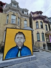 A large artwork of a man's portrait with a yellow background and a blue coat is displayed outdoors. The portrait is positioned in front of a row of Victorian-style brick houses with ornate architectural details.