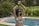 A person wearing sunglasses and a bikini top holds a paddle or racket above their head. They are standing next to a swimming pool in a lush, green garden. In the background, there are some trees, bushes, and outdoor lounge chairs.