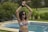 A person wearing sunglasses and a bikini top holds a paddle or racket above their head. They are standing next to a swimming pool in a lush, green garden. In the background, there are some trees, bushes, and outdoor lounge chairs.