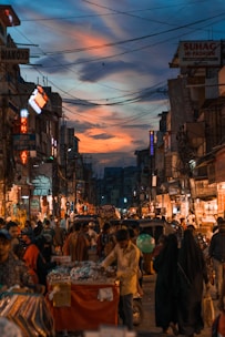 A bustling street market with numerous vendors, shops, and signs lining both sides of the street. People are walking around, interacting, and shopping. The sky above is painted with dramatic colors of orange, blue, and pink, suggesting the time is around sunset. Overhead wires crisscross the scene, adding to the urban feel.