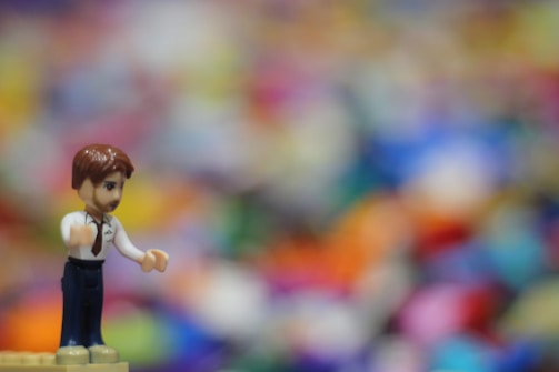 A small toy figure resembling a human, featuring a brown-haired character in a white shirt with a tie and dark pants, stands sharply in focus against a colorful, blurry background. The multitude of colors in the background creates a sense of whimsy and abstraction.