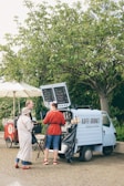 A small coffee cart set up in a converted vehicle is parked under a tree. A person in a red sweater and jeans is serving two customers at the counter. The surroundings are lush with greenery, including another cart and umbrellas nearby.
