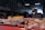 A market stall displays various cuts of raw meat on trays and a wooden block inside a dimly lit warehouse. In the background, a vendor wearing a red apron with a grey shirt is somewhat blurred, possibly preparing or selling the meat.