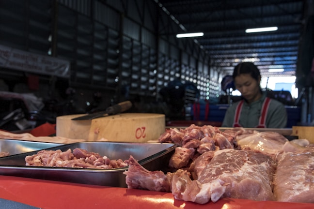 A market stall displays various cuts of raw meat on trays and a wooden block inside a dimly lit warehouse. In the background, a vendor wearing a red apron with a grey shirt is somewhat blurred, possibly preparing or selling the meat.