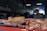 A market stall displays various cuts of raw meat on trays and a wooden block inside a dimly lit warehouse. In the background, a vendor wearing a red apron with a grey shirt is somewhat blurred, possibly preparing or selling the meat.