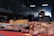 A market stall displays various cuts of raw meat on trays and a wooden block inside a dimly lit warehouse. In the background, a vendor wearing a red apron with a grey shirt is somewhat blurred, possibly preparing or selling the meat.