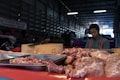 A market stall displays various cuts of raw meat on trays and a wooden block inside a dimly lit warehouse. In the background, a vendor wearing a red apron with a grey shirt is somewhat blurred, possibly preparing or selling the meat.