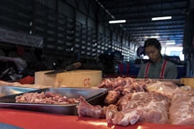A market stall displays various cuts of raw meat on trays and a wooden block inside a dimly lit warehouse. In the background, a vendor wearing a red apron with a grey shirt is somewhat blurred, possibly preparing or selling the meat.