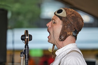 A man wearing a vintage leather aviator helmet and white shirt is speaking or singing into a retro-style microphone. The background is slightly blurred with a mix of indoor and outdoor elements.