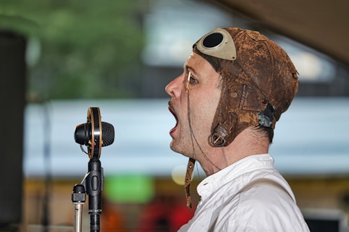 A man wearing a vintage leather aviator helmet and white shirt is speaking or singing into a retro-style microphone. The background is slightly blurred with a mix of indoor and outdoor elements.