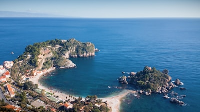 An aerial view of a coastal landscape featuring two rocky islands covered in greenery. The larger island is connected to a mainland area with buildings and a beach. The calm blue sea surrounds the islands, creating a serene and picturesque scene.
