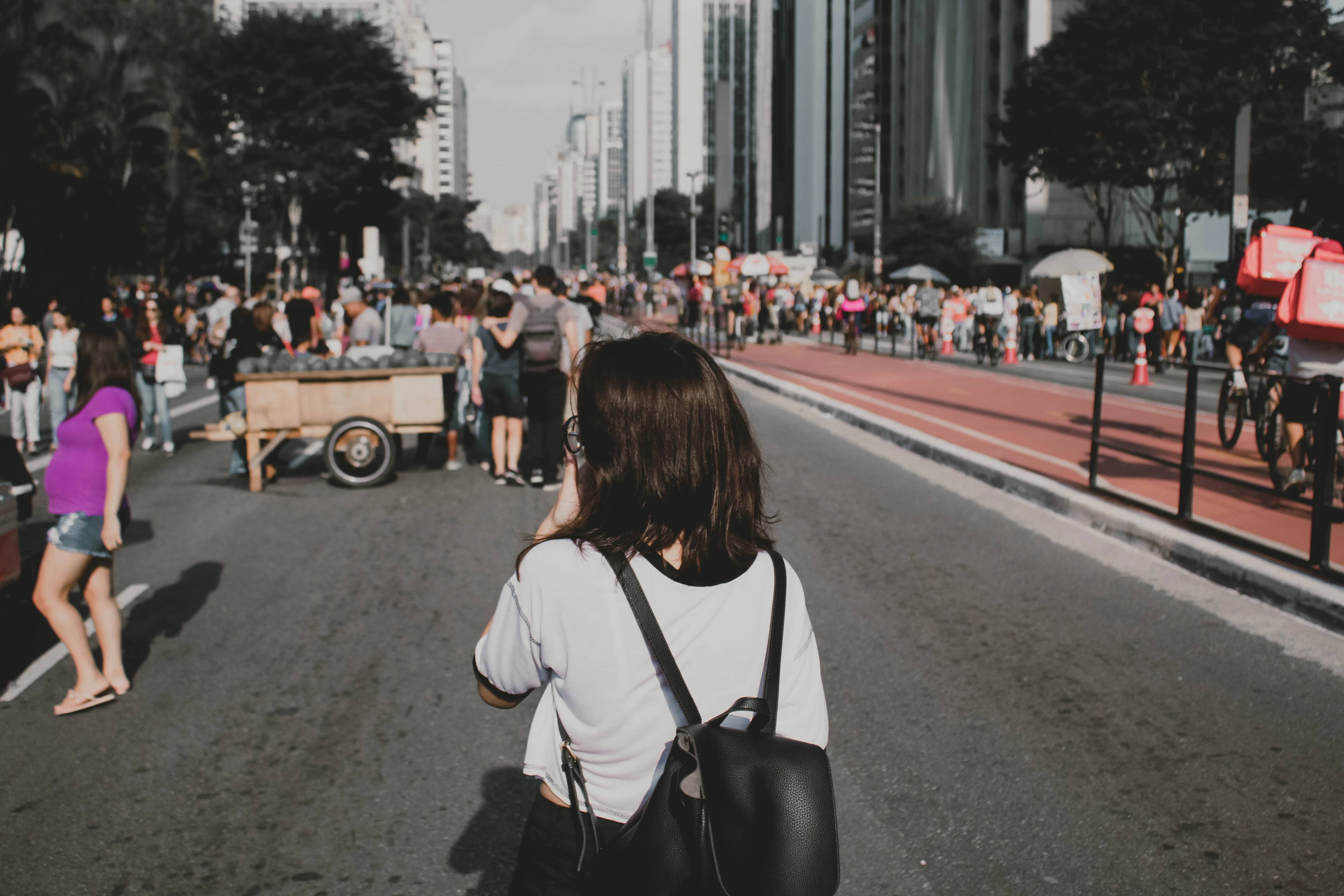 A woman walks down the wide lanes of Avenida Paulista past a food cart and a crowd on a sunny afternoon.