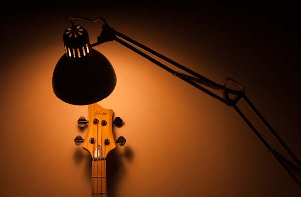 A black desk lamp illuminates the upper part of a guitar neck against a warm, neutral background. The light casts dramatic shadows, creating a cozy and intimate atmosphere.