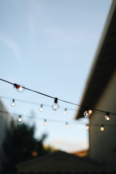 A string of round bulb lights is suspended across an outdoor area in the late afternoon or early evening. The sky is clear and light blue, and the silhouette of a building is visible on one side. The lighting creates a warm, inviting atmosphere.