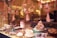 A display window showcases a variety of beautifully decorated cakes and pastries arranged on different tiers. The backdrop features soft, floral decorations hanging above the desserts. The ambient lighting casts a warm glow, highlighting the intricate details of the baked goods.