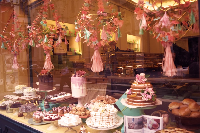 A display window showcases a variety of beautifully decorated cakes and pastries arranged on different tiers. The backdrop features soft, floral decorations hanging above the desserts. The ambient lighting casts a warm glow, highlighting the intricate details of the baked goods.