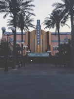 A retro-styled theater with a prominent Hollywood sign is flanked by tall palm trees. The facade features art deco architectural elements and a notable marquee displaying movie showtimes. Street lamps with orb lights line the entrance, creating a classic cinematic atmosphere.