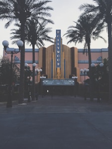 A retro-styled theater with a prominent Hollywood sign is flanked by tall palm trees. The facade features art deco architectural elements and a notable marquee displaying movie showtimes. Street lamps with orb lights line the entrance, creating a classic cinematic atmosphere.