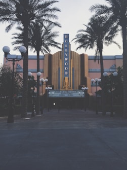 A retro-styled theater with a prominent Hollywood sign is flanked by tall palm trees. The facade features art deco architectural elements and a notable marquee displaying movie showtimes. Street lamps with orb lights line the entrance, creating a classic cinematic atmosphere.