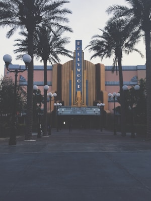 A retro-styled theater with a prominent Hollywood sign is flanked by tall palm trees. The facade features art deco architectural elements and a notable marquee displaying movie showtimes. Street lamps with orb lights line the entrance, creating a classic cinematic atmosphere.