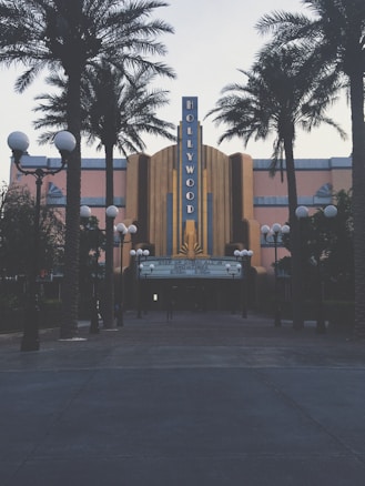 A retro-styled theater with a prominent Hollywood sign is flanked by tall palm trees. The facade features art deco architectural elements and a notable marquee displaying movie showtimes. Street lamps with orb lights line the entrance, creating a classic cinematic atmosphere.