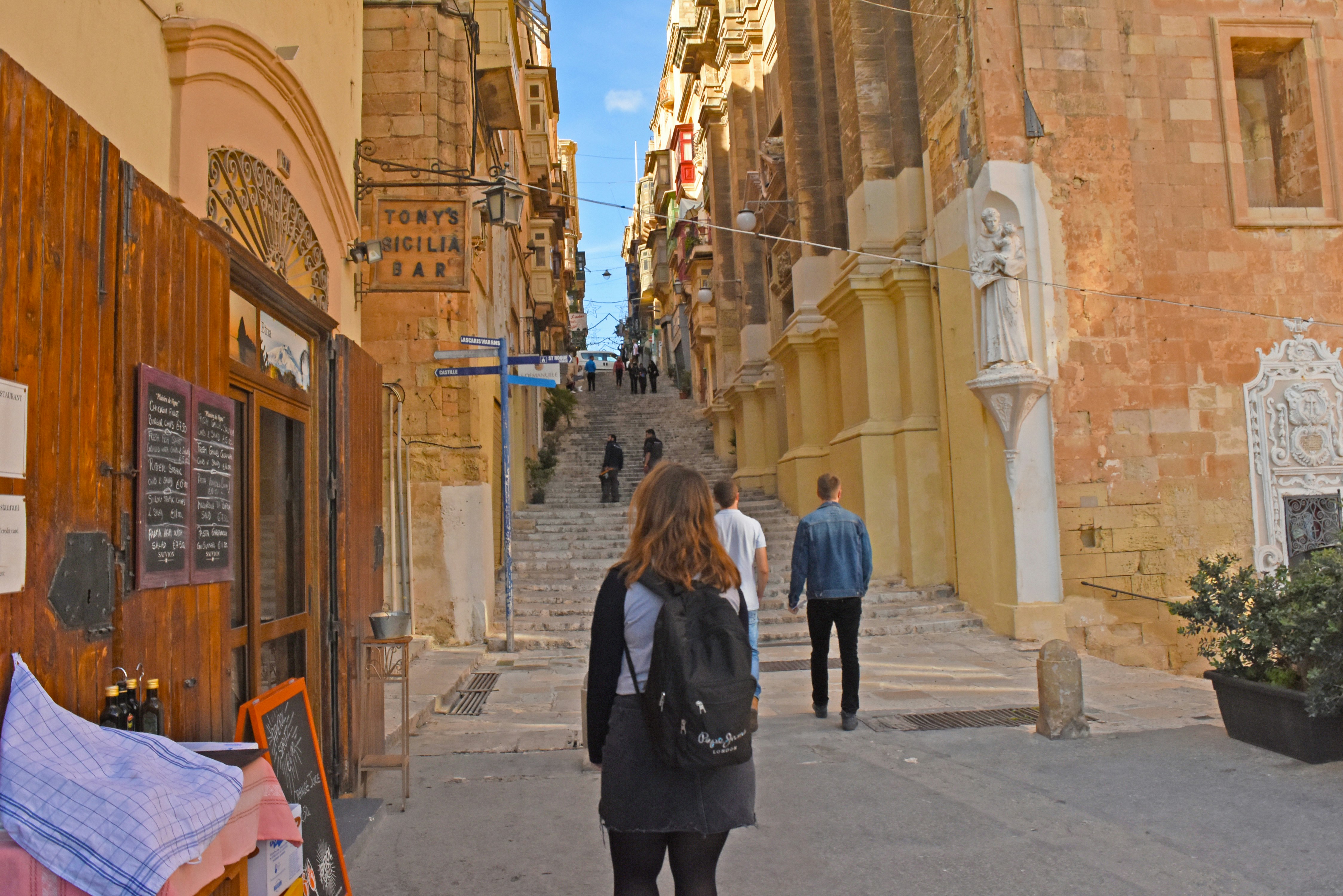 woman in black coat standing on sidewalk during daytime, Calles antiguas de Malta.
