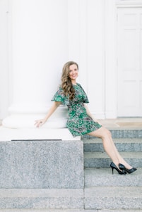 A woman with long, wavy hair is seated on stone steps near a large white column. She is wearing a green dress with a pattern and black high-heeled shoes. The background features a white paneled door, contributing to the bright and airy setting.