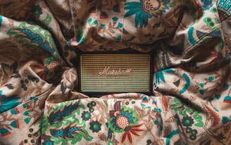 A vintage-style speaker rests on a fabric with intricate floral and leaf patterns. The speaker has a woven front with a logo in the center, surrounded by rich colors from the fabric including greens, reds, and blues. The scene appears cozy and artistic.