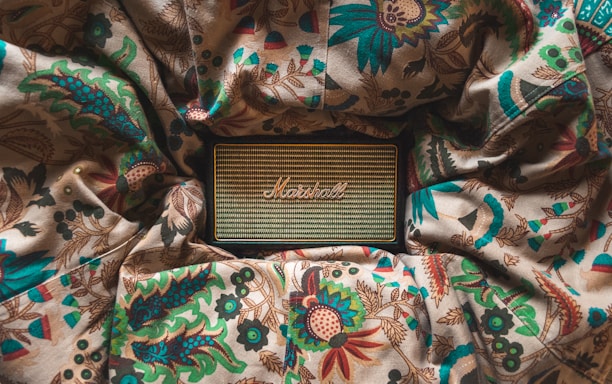 A vintage-style speaker rests on a fabric with intricate floral and leaf patterns. The speaker has a woven front with a logo in the center, surrounded by rich colors from the fabric including greens, reds, and blues. The scene appears cozy and artistic.