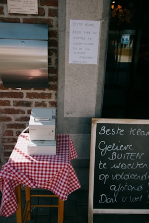 A small table covered with a red and white checkered tablecloth holds an Epson printer. Next to it, a chalkboard with handwritten text in Dutch advises customers to wait outside after taking a number. The scene includes a brick wall and a metal mailbox.