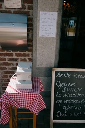 A small table covered with a red and white checkered tablecloth holds an Epson printer. Next to it, a chalkboard with handwritten text in Dutch advises customers to wait outside after taking a number. The scene includes a brick wall and a metal mailbox.