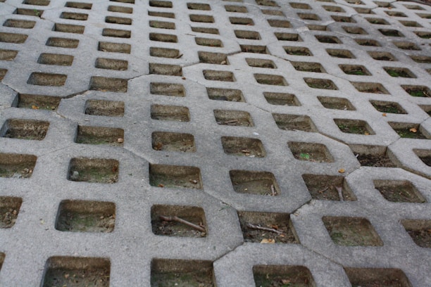 A concrete surface with a pattern of square holes arranged in a grid. The holes reveal patches of earth and some scattered twigs and small debris. Cracks are visible in the concrete, adding texture to the surface.