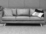 A three-seat leather couch with a mid-century modern design, adorned with patterned throw pillows. The couch is placed on a textured rug against a backdrop of a tiled wall. The overall setting appears minimalist and stylish.