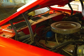 A detailed view of a car engine with a red hood partially open. The air filter is prominently displayed, and various engine components and hoses are visible, suggesting a high-performance setup.